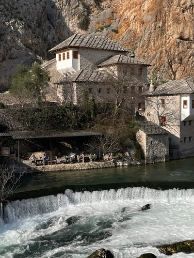 The image depicts a stone architectural complex situated at the base of a large, rugged cliff face in Dračevice, Bosnia and Herzegovina, likely during daylight hours. In the background, a massive, multi-toned cliff, primarily orange-brown with gray sections, rises steeply. The middle ground features several traditional Ottoman-style stone buildings with pitched roofs covered in stone tiles. One prominent building exhibits a white, projecting bay window with three narrow wooden-framed windows. Sparse, bare-branched trees and green foliage, including ivy, are distributed around the buildings and along the lower cliff areas.
In the foreground, a greenish-blue river flows from right to left, interrupted by a wide, foamy waterfall or weir spanning its width. Below the waterfall, the water is turbulent and white. Along the riverbank on the left, an outdoor restaurant area is visible, sheltered by a dark, corrugated roof supported by wooden posts. This area features multiple tables and chairs, with approximately 6-8 individuals seated, indistinctly visible due to distance and shadow, engaged in dining or leisure. Potted plants are present on some tables. The scene is illuminated by strong sunlight, casting distinct shadows from trees onto the building walls and highlighting textures on the cliff and water.