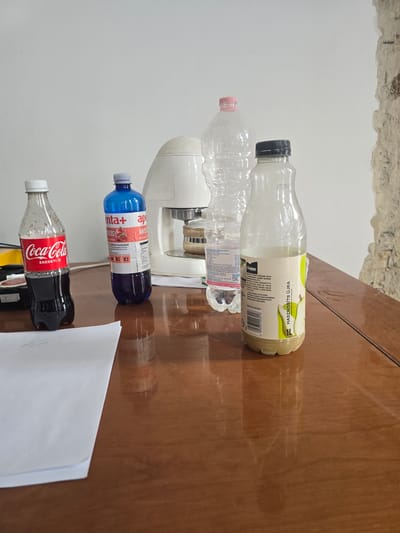 The scene is an indoor setting on a polished wooden table in Nyíregyháza, Hungary. Five plastic beverage bottles are arranged on the table from left to right: a partially full 1.2-liter Coca-Cola bottle with a white cap; a mostly full blue bottle of "enta+ ape ANTIOXPRESS" with a grey cap; an empty 1.5-liter clear plastic bottle with a pink cap; a partially full 1.5-liter "NaturaQUA" branded clear plastic water bottle with a black cap; and a partially full 1-liter Cappy pear juice bottle with a black cap. A white kitchen appliance with a metallic base, possibly a coffee maker, stands behind the central bottles. In the foreground, a stack of white paper sheets with faint, illegible handwriting rests on the table. The background features a plain white wall on the left, transitioning to an exposed, rough, light-colored wall section, possibly concrete or brick, on the right. No individuals are present, and no actions or interactions are depicted.