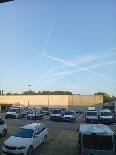 The image shows a parking lot in Strengelbach, Switzerland. The lot is filled with various vehicles, including several white vans, a white station wagon, and a truck. In the background, there is a large, beige building with a grid pattern. Above the building, the sky is a clear blue with several white contrails from airplanes forming an "X" shape. The sun is casting long shadows, suggesting it is either early morning or late afternoon. The overall scene depicts a typical day in a commercial or industrial area.