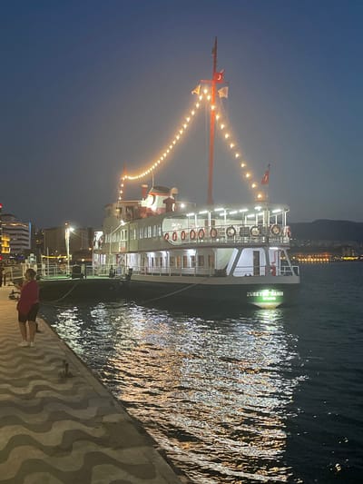 The image shows a ferry docked at a pier in Izmir, Turkey, at dusk. The ferry, named "ZÜBEYDE HANIM ISTANBUL," is illuminated by string lights that create a warm glow across its deck. The ferry is white with red accents and has multiple decks, with life preservers visible along the railing. The water around the ferry reflects the lights of the city and the ferry itself. On the left side of the image, a person stands on the pier, looking towards the water. The pier has a distinctive wavy pattern. In the background, buildings and distant hills are visible, with more city lights suggesting a bustling urban environment. The sky is a deep blue, indicative of the twilight hour.
