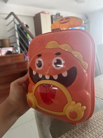A person holds an orange plastic container with a cartoon hamburger design. The container features a yellow lid with a "HAMBURGER" label. The background includes a living room or dining area, with furniture like shelves and a table visible. The specific location is La Victoria, Peru, as per the prompt.