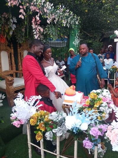 This image captures an outdoor wedding celebration in Amokvo, Nigeria. The central focus is a dark-skinned male groom, wearing a red blazer with black lapels and a black shirt, standing alongside a dark-skinned bride. The bride wears a white strapless wedding gown with a full skirt, a prominent silver necklace, and earrings; her black hair is pulled back. Both individuals are smiling.
The couple is in the process of cutting a two-tiered wedding cake with a shared knife. The cake, resting on a small wooden stand, features a peach-orange top tier with a rounded top and red trim, and a white bottom tier with an orange band. A young dark-skinned male child, dressed in a red blazer matching the groom's, stands between the couple, facing the cake, with his right hand near the knife.
To the right of the couple, an adult dark-skinned male, wearing a teal-blue traditional Nigerian garment with embroidered details and a black messenger bag, holds a black microphone in his right hand, addressing the gathering. Behind him, numerous adults and children are visible as spectators.
The ground is covered with green artificial turf. The backdrop includes elaborate decorations: artificial green and light-pink foliage hangs from an overhead structure, creating a canopy effect. Ornate gold-colored chairs with white upholstered seats are positioned to the left. A white decorative barrier in the foreground is adorned with numerous artificial flower bouquets in gold, silver, pink, purple, light blue, and white. Natural trees and dense foliage are visible in the distant background.