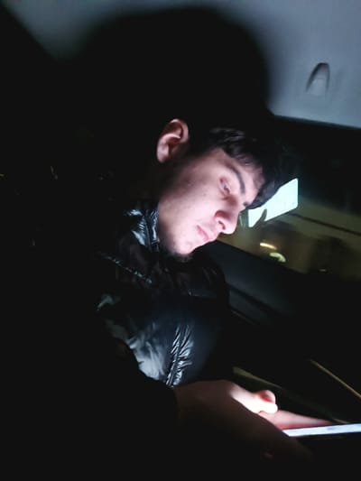 A young man sits in a dark car, illuminated by the light of a phone he is holding. He wears a puffy black jacket. His gaze is directed downwards at the phone. The interior of the car is barely visible, showing a headrest, window, and faint light from outside. The overall scene is dimly lit.