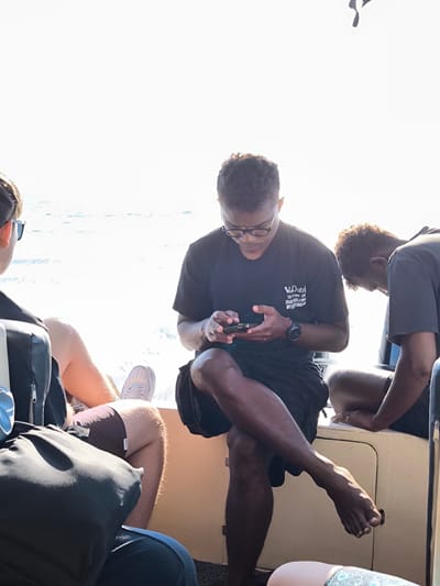 The image shows three people sitting on a boat, possibly on a tour in Pasir Putih, Indonesia. The person in the center, wearing glasses and a black t-shirt, is looking down at their smartphone. Their legs are crossed, and they are wearing shorts. To their left, another person is partially visible, wearing sunglasses and shorts. To the right, a third person is hunched over, also looking down. The background shows the bright sky and the shimmering water, indicating a sunny day. The boat appears to be a simple passenger vessel.