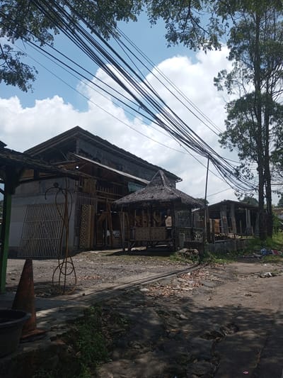 The image shows a street scene in a cluttered, possibly underdeveloped area. A large building with a partially visible sign and what appears to be bamboo construction materials and finished products is in the background. There is a small, thatched-roof structure in front of the building. Power lines are strung across the sky. A traffic cone and black bucket sit at the edge of the road. The road surface is uneven and littered with debris. Trees are visible on either side of the frame. The location is purported to be in Kioumaknao 2, Laos. No people are visible.