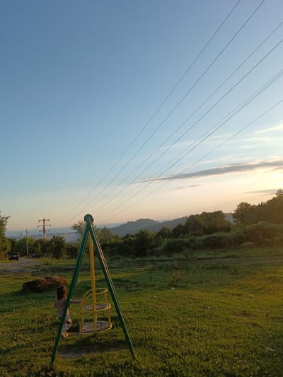 This is a medium shot, outdoor, eye-level, vertical, natural light photograph taken in Chereshnya, Russia. The scene depicts a child standing by a swing set in a grassy field under a clear blue sky. The swing set is green and yellow, with a child facing away from the camera, touching the swing. A car is parked on a dirt road to the left of the frame, near a large rock. In the background, power lines stretch across the sky, and a hill covered in trees is visible. A power line tower with red and white markings stands in the distance. The sun is setting, casting a warm glow over the landscape, with soft clouds visible near the horizon.