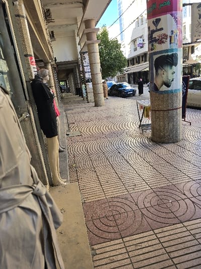 This image depicts an urban street scene in Taza, Morocco, under bright daylight. The foreground on the left shows the back of a mannequin dressed in a beige trench coat, tied at the waist. To its right, another mannequin, partially visible behind a storefront, wears a black top and displays bare, light-toned legs.

The sidewalk, a central feature, is paved with distinct patterns: reddish-brown tiles with concentric circles interspersed with lighter-toned tiles featuring linear and curvilinear designs. Several multi-segment concrete columns support an overhead concrete structure, forming a covered walkway. The closest column on the right is heavily adorned with various posters, including a prominent advertisement for a barber shop featuring a man with a modern haircut, and other posters with Arabic text and product imagery. A small, light-colored display stand with a draped fabric is situated adjacent to this column.

Further into the midground, a paved street runs parallel to the sidewalk. A dark-colored sedan is parked on this street. Two individuals are present: one person, dressed in a light-brown traditional Moroccan garment (Djellaba), stands near the parked car. Another person, clad in dark clothing and a head covering, walks away from the camera further down the street.

The background reveals a series of multi-story buildings lining the street. A tall, modern building with reflective windows is visible in the distance. A green tree provides foliage, and overhead utility lines traverse the scene.