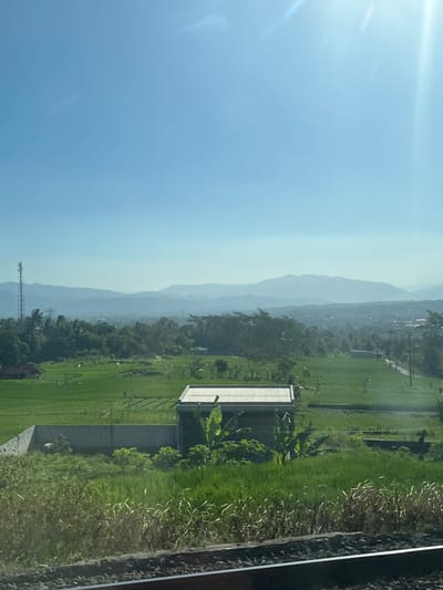 The image depicts a rural landscape under a clear blue sky with bright sunlight creating lens flares. In the foreground, train tracks run horizontally across the bottom of the frame. Beyond the tracks, a vibrant green field is visible, with some dry, tall grass in the immediate foreground. Further into the midground, a rectangular building with a corrugated metal roof stands amidst lush greenery, possibly banana trees. Adjacent to this building is a concrete wall, behind which a person is partially visible, appearing to be engaged in some activity. Beyond this, more terraced green fields, likely rice paddies, stretch out, dotted with small structures and trees. In the background, a hazy line of mountains forms the horizon, and a tall telecommunications tower is visible to the left of the frame. The overall scene suggests a serene countryside setting in the region of Negaradaha, Indonesia, characterized by agricultural land and natural topography.