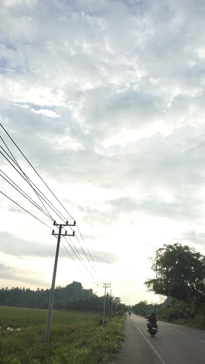 An overcast sky looms above a rural Indonesian landscape in the city of Cotentung. A paved road stretches into the distance, flanked by lush green vegetation and rice paddies. Several power lines crisscross the frame, supported by utility poles. In the foreground, a single utility pole stands prominently on the left side of the road. Further down the road, two motorcycles are visible, one with a rider and another in the distance. A large tree with dense foliage is situated on the right side of the road. The overall scene depicts a typical day in a rural Indonesian setting.