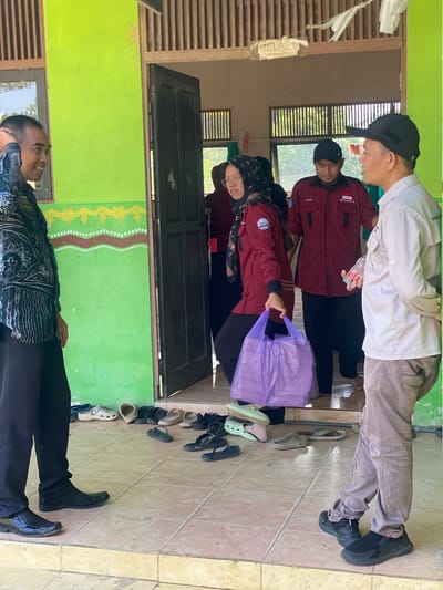 In Handil Baru, Indonesia, a group of individuals stands on a tiled porch outside a building with a lime green exterior and a wooden doorframe. A sign above the doorway reads "KELAS 6," indicating it is a classroom. Four people are visible. On the left, a man in a dark patterned shirt and black pants smiles and touches his head, facing two people entering the doorway. Inside the doorway, a woman in a maroon jacket, headscarf, and glasses holds a large purple plastic bag filled with items. To her right, a man in a maroon jacket with the Indonesian flag and text on it, and another man in a light-colored shirt and brown pants are visible. The man in the maroon jacket is looking down, and the man in the light-colored shirt is speaking to the man on the left. Various shoes, including sandals and crocs, are neatly arranged on the floor near the entrance, suggesting a practice of removing footwear before entering. The environment appears to be outdoors, with sunlight casting shadows on the tiled floor.