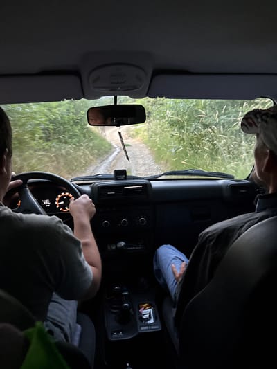 The image shows the interior of a car driving on a dirt road in a rural area, likely Lavrovoe, Ukraine. The perspective is from the back seat, looking forward. Two individuals are visible. The driver, in the foreground left, is wearing a grey t-shirt and has their hands on the steering wheel. The dashboard displays illuminated instruments, including a speedometer and what appears to be an odometer reading "1127". To the right of the driver, a passenger wearing a baseball cap and a dark jacket is seated. The passenger's legs are visible, clad in blue jeans. The exterior environment, seen through the windshield and side windows, consists of dense green foliage and tall grasses bordering the narrow dirt road. A rearview mirror hangs from the windshield, reflecting a partial view of the driver's face and the road ahead. A small black air freshener is attached to the dashboard. The overall scene depicts a journey through a natural, unpaved environment.
