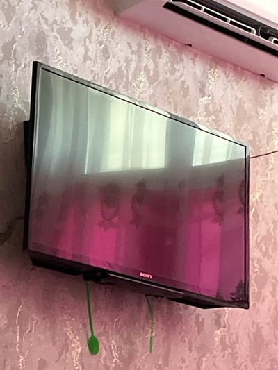 A flat-screen television is mounted on a textured pink wall in Türkistan qalası, Kazakhstan. The TV screen reflects a window with vertical blinds and a pink curtain. The SONY logo is visible on the bottom left corner of the TV. Two green cables hang down from the bottom of the television. An air conditioning unit is mounted above the television. The overall scene suggests a domestic or commercial interior setting.
