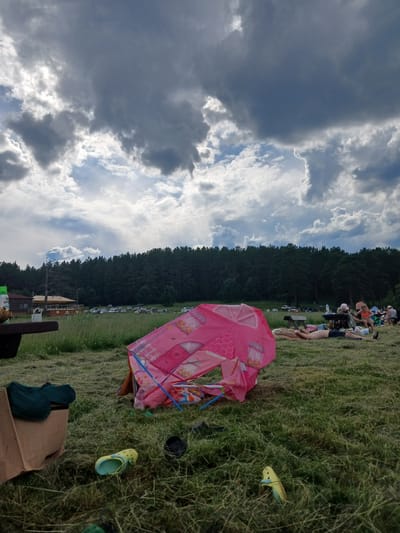 The image depicts an outdoor gathering at a grassy area with a dense forest in the background. In the foreground, a pink children's tent is partially collapsed. Various items are scattered on the grass, including a green item of clothing in a cardboard box, a yellow Croc shoe, and a black shoe. Several people are visible in the midground, some lying down and others sitting. They appear to be picnicking or relaxing. A line of cars is parked in front of the forest, suggesting a public park or recreational area. The sky is filled with dramatic, dark clouds, indicating potential for rain. The location is identified as the city of Revda, Russia.