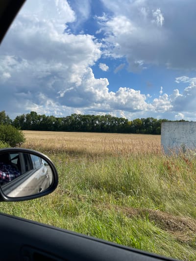 The image is a photograph taken from inside a car, looking out the passenger side window towards a rural landscape in Stremyhorod, Ukraine. The foreground shows the interior of the car door and the side-view mirror, which reflects a person wearing a red, blue, and white plaid shirt. Beyond the car, a field of dry, golden grass stretches towards a line of lush green trees that form a dense forest. The sky is a vibrant blue, dotted with dramatic, puffy white clouds. To the right of the frame, a portion of a weathered, light-colored concrete structure is visible, partially obscuring the field. The overall scene depicts a sunny day in the Ukrainian countryside.