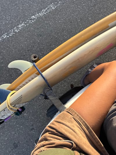 In Denuwala, Sri Lanka, three surfboards are being transported, secured to a vehicle by a blue bungee cord and straps. The surfboards are layered diagonally, with the top board appearing to be a bright yellow or orange, the middle board a lighter cream color with pink markings, and the bottom board a similar cream color with black lines. A light-colored fin with the brand "SHAPER" is visible on the middle surfboard. A person's legs, tanned and wearing brown shorts, are positioned in the foreground, suggesting they are either the operator or passenger of the vehicle. The surfboards are positioned over a dark, possibly black, object, which could be part of the vehicle's rack or frame. The background consists of dark asphalt with a faint white line, indicating a road surface. The lighting suggests it is daytime, with shadows cast on the road.