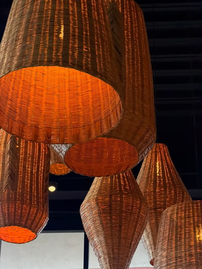A cluster of approximately eight woven light fixtures, appearing to be crafted from wicker or rattan, hangs from a dark ceiling within an indoor setting in Fraccionamiento Los Heroes Queretaro, Mexico. The fixtures exhibit various shapes, including truncated cones, cylinders, and elongated teardrop or bell forms, with their open ends facing downwards or angled slightly. Each fixture contains an internal light source, emitting a warm, amber-orange glow that radiates through the woven material and brightly illuminates the interior surfaces. The light casts visible striations and patterns due to the weave. The background consists primarily of a dark, uniform ceiling, with hints of lighter, out-of-focus architectural elements, possibly walls or windows, visible towards the lower left and center-right of the frame. One small, unlit recessed downlight is visible on the dark ceiling in the upper mid-left quadrant. No individuals or specific actions are depicted.