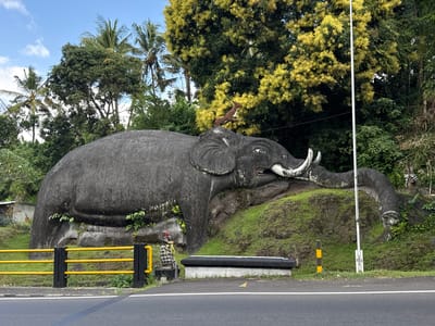 The image depicts a large concrete elephant statue situated beside a road in Banjarangkan, Indonesia. The elephant is positioned on a grassy embankment, with its trunk extended and head facing right. A small, spotted animal statue is perched on the elephant's head. In front of the elephant, a yellow metal barrier separates the road from the embankment. A small shrine with a figure draped in checkered fabric is visible to the left of a black stone bench. The background features lush green trees, some with yellow flowers, and a blue sky with scattered clouds. A white pole, possibly a street light or utility pole, stands to the right of the elephant. The road in the foreground is asphalt with white markings.