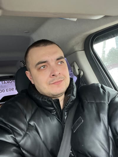 A male individual is observed seated inside the passenger compartment of a vehicle in Rogachovo, Russia. He appears to be an adult, exhibiting short dark hair, light brown eyes, and light facial stubble. His gaze is directed upwards and to the right, away from the camera, with a neutral expression and a slight upturn at the corners of his mouth. He is attired in a black, shiny, puffer-style jacket, secured by a visible black seatbelt diagonally across his torso and left shoulder. The vehicle's interior features a light gray headliner and trim. Through the passenger window on the right, a blurry outdoor scene consistent with overcast or winter weather is visible, showing indistinct trees or foliage. On the left side of the frame, partially obscured and reflected, is Cyrillic text, appearing inverted as "ТАКОР" (likely part of "ПРОКАТ" - Rental) and numbers "000 ε0ε" (likely "303 000"), suggesting an external commercial establishment. The scene depicts the individual in a stationary or moving vehicle.