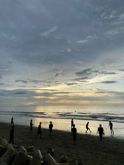 The image depicts a group of individuals playing soccer on a beach at dusk in Bateetimoh, Indonesia. The sky is filled with dramatic clouds, with a warm glow of sunset visible on the horizon over the calm ocean. The figures of the players are silhouetted against the fading light as they interact on the wet sand near the water's edge. In the foreground, large concrete blocks, likely part of coastal defense structures, are visible on the beach. The overall atmosphere is serene and captures a moment of leisure and sport in a natural coastal setting.