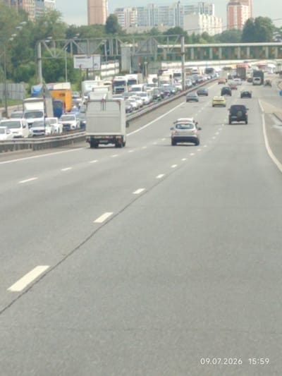 The image shows a traffic jam on a multi-lane highway with buildings and trees in the background. The road is asphalt, with white lane markings. There are numerous vehicles on the highway, including cars, trucks, and vans, all moving slowly or stopped. The sky is overcast. A timestamp in the lower right corner indicates the date and time as 09.07.2026 15:59. The location is identified as Vinogradovo, Russia.