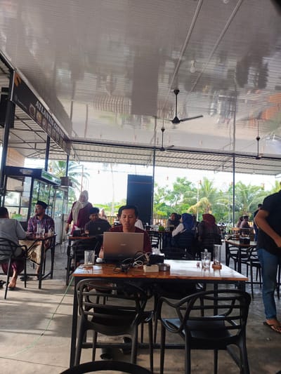 This is a medium shot of an outdoor cafe in Peurupok, Indonesia, likely during the daytime. The scene is filled with people seated at tables, engaged in various activities. In the foreground, a man is engrossed in working on his laptop, with a drink and cables on the table. Several other individuals are visible at tables in the mid-ground and background, some conversing, others working, and one woman appears to be walking between tables.
The cafe has a covered roof supported by pillars, with ceiling fans for air circulation. Palm trees and lush greenery are visible in the background, suggesting a tropical or sub-tropical climate. The flooring appears to be concrete. The overall atmosphere seems relaxed and casual, typical of a cafe setting.
A banner above some of the tables reads "ATLANTIS COFFEE" and displays images of food items. There are also reflections visible on the shiny ceiling, showing glimpses of the surroundings. The presence of laptops, drinks, and the general layout of the cafe indicate it's a place for people to gather, work, or socialize. The lighting suggests it's daytime, but the exact time is not specified.