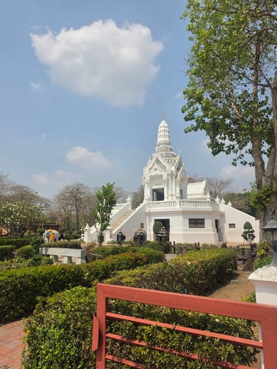 The image depicts a white temple complex in the Historic City of Ayutthaya, Thailand. The temple is multi-tiered with ornate detailing. It is flanked by landscaped gardens with trimmed hedges. A tree with green leaves overhangs the left side of the frame. In the foreground, a red metal fence is visible. The sky is blue with scattered white clouds.
