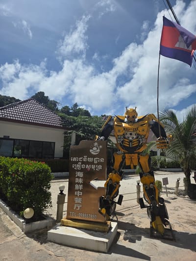In Koh Toch Village, Cambodia, a large, yellow, and black Bumblebee robot sculpture stands prominently on a sandy ground, casting a long shadow. The robot is positioned in front of a golden sign with Asian script and a phone number, "TEL: 098667890," and what appears to be a seafood restaurant logo. To the left of the robot, a white building with a dark tiled roof is visible, bordered by lush green bushes. The sky above is a bright blue with scattered white clouds. A Cambodian flag flutters from a pole to the right of the robot. Palm trees are present in the background, suggesting a tropical or coastal environment.