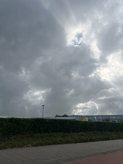 This is a vertical, outdoor, eye-level shot of a cloudy sky above a modern building, a tall hedge, and a paved walkway. The sky is filled with thick, gray clouds, with some patches of lighter sky and sunbeams breaking through on the left side of the frame. A single streetlamp is visible in the upper right portion of the frame, extending from the right.
The building, visible on the left, appears to be multi-story with a facade of windows. Below the building is a dense, dark green hedge that stretches horizontally across the middle ground. To the right of the hedge is a paved walkway made of dark gray rectangular stones, with a reddish-brown border along the far right edge.
The scene suggests it is daytime, likely overcast or transitioning weather, given the heavy cloud cover and visible sunbeams. The environment appears urban or suburban, possibly a park or a commercial area, given the building and paved walkway. There are no people or animals visible in the frame. The overall mood is somewhat subdued due to the cloudy weather. There is no visible text in the image.