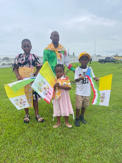 Four people are holding flags on a grassy lawn.

A young boy is in the top center wearing a gold baseball cap, a white shirt with a graphic, and tan shorts. He is holding a flag of the Vatican City and a flag of Equatorial Guinea.

Below the boy, an older person with a shaved head is wearing a green and gold patterned shirt with a scarf of similar material.

Below the older person, a young girl is wearing a dress with a pink skirt and a top of the same pattern as the older person's shirt. She is holding a flag of the Vatican City.

Below the young girl, a young person is wearing a black floral patterned shirt and black shorts with gold patterning. They are also holding a flag of the Vatican City.

The location is Malabo, Equatorial Guinea. A fence can be seen in the background, along with a black SUV. The sky is overcast.