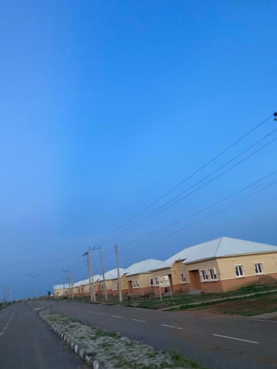 The image captures a residential street in Dumnga, Nigeria, with a row of light-colored, single-story houses lining the right side of a paved road. The houses have cream-colored walls with a darker, possibly terracotta-colored lower section, and white roofs. They feature numerous windows with white frames. Overhead, power lines stretch across the clear blue sky, supported by utility poles. The road has white lane markings, and the verge is lined with a curb and sparse green vegetation. The scene appears to be set during daylight hours, with the clear sky suggesting fair weather. There are no people visible in the frame.
