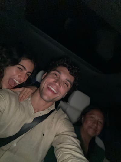 Three smiling individuals pose in what appears to be the backseat of a car. A man with dark, curly hair and a light button-down shirt is in the center. A woman with dark, curly hair is to his lower left, mouth open in a wide grin. A third individual is in the upper right, peering towards the camera. The lighting is dim, with a large area of black in the upper left. A headrest is visible behind the central figure.