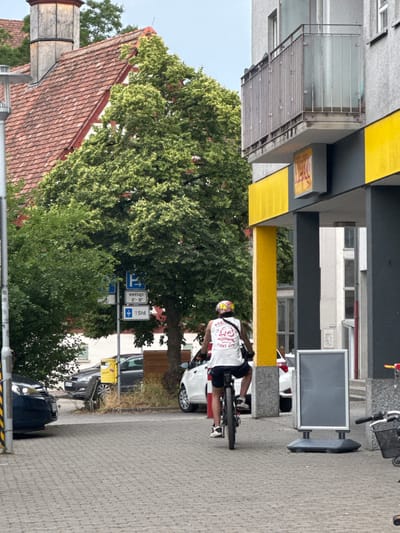 In Sindelfingen, Germany, on a cobblestone pedestrian area, a person wearing a white t-shirt with a graphic design, black shorts, and a helmet with goggles is riding a bicycle away from the camera. The t-shirt has the text "BOXING EST 1994" and "AESTHETICS FIGHT GYM". The bicycle is dark colored with visible spokes on the rear wheel. To the right of the cyclist, a tall, rectangular A-frame sign with a blank gray display is situated on a black base. Further to the right, a portion of a bicycle with a basket and a red accessory is visible. In the background, a multi-story building with a gray facade and balconies with metal railings stands. Yellow and dark gray panels are prominent features of the building's lower level. Adjacent to the building is a large tree with green foliage. To the left of the tree, a blue parking sign with white symbols and text indicating "werktags 8-18" and "1 Std." is mounted on a pole. Several cars are parked in the vicinity, including a dark-colored car on the left and white cars further back. A red-tiled roof of an older building is visible behind the tree on the left, topped with a weathered chimney structure. The sky is overcast.