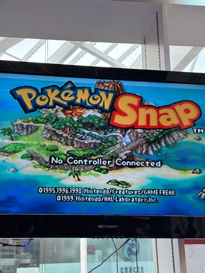 An image displays the title screen of the video game "Pokémon Snap" on a television. The game's title, "Pokémon Snap," is prominently featured in large, colorful text across the top portion of the screen. Below the title, a vibrant, tropical island landscape is depicted, complete with lush greenery, sandy beaches, and clear blue water. A winding path is visible on the island, suggesting a gameplay element. Centered on the screen, in white text, is the message "No Controller Connected," indicating the game is paused or awaiting input. At the bottom of the screen, copyright information is visible, listing "©1995, 1996, 1998 Nintendo/Creatures/GAMEFREAK" and "©1999 Nintendo/HAL Laboratory, Inc." The television itself is black and appears to be an older model, with visible buttons and branding on its lower bezel that reads "Emerson." The television is positioned in front of a window that allows natural light to enter, illuminating the scene. Through the window, the ceiling structure of what appears to be a commercial space, possibly a store or exhibition hall, is visible. The image seems to have been taken in El Paso, United States of America.