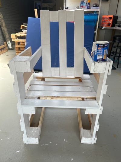 The image shows a white-painted chair made from wooden pallets, placed on a concrete floor in what appears to be a garage or workshop. The chair is in the foreground and is the primary subject. A can of "Spectrum Gloss paint" in white is visible on the armrest of the chair. In the background, there are shelves with various items, including bins and cleaning supplies, suggesting a storage or workspace. A large blue sheet or mat is leaning against the wall behind the chair. The overall scene is cluttered and industrial, consistent with a DIY project or a workspace. The image does not provide any information to suggest this location is in Lisse, Netherlands.