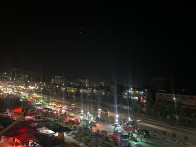 This is a night-time aerial view of a bustling street market in Kabul, Afghanistan. The scene is illuminated by a variety of lights from street lamps, market stalls, and passing vehicles. 

In the foreground, numerous market stalls are set up, covered with colorful tarps, primarily red and green. Some stalls appear to be selling produce, with baskets of goods visible. People are actively engaged in commerce, with several individuals seen walking through the market and interacting with vendors. Small carts and hand-pulled vehicles are also present, likely used for transporting goods.

The street in the midground is filled with traffic, including cars, motorcycles, and what appear to be auto-rickshaws. Headlights and taillights create streaks of light, indicating movement. On the far side of the street, several buildings are visible, some multi-story, with illuminated windows and storefronts. Trees line the street and the area around the buildings.

In the background, the city skyline is visible against the dark night sky. There are more buildings, some taller than others, and faint lights suggest further urban development. The sky itself is mostly dark, with a few scattered pinpricks of light, possibly stars or distant lights. The overall impression is of a vibrant, active urban environment at night.