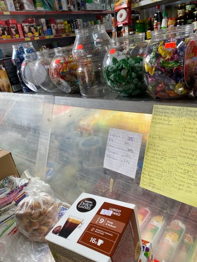 This image captures an interior view of a small retail store or grocery shop in Sredkovets, Bulgaria. The perspective is from slightly above a counter, looking into the establishment.

In the foreground, on the left, a brown cardboard box with the word "OPEN" partially visible is present. To its right, a clear plastic bag contains numerous small, light brown, rounded objects exhibiting sprouts, consistent with onions or small potatoes. A Nescafé Dolce Gusto Lungo Intenso coffee capsule box, displaying a price sticker of "5.99 лв" (Bulgarian Leva), is prominently placed on the counter. The box specifies "Intensity 9 Deep & Robust" and contains "16x" capsules.

Behind the counter, separated by a clear barrier, is a display area featuring a row of large, clear plastic jars. These jars are filled with various confections; one contains uniformly green-wrapped candies labeled in Cyrillic as "лyлче дюлякови," and another holds an assortment of colorful, individually wrapped candies, including "Toffy" and "Jelly" brands. Several other similar jars, some partially empty, are also present.

Two handwritten notes in Cyrillic are taped to the surface behind the counter. A white note lists items and prices such as "Хляб - 4.55 лв" (Bread - 4.55 BGN) and "Колбаси - 1.6 лв" (Sausages - 1.6 BGN). A larger yellow note to the right presents an extensive list of products, including various types of "Пиво" (Beer), with corresponding prices.

The background reveals multiple retail shelves densely stocked with a diverse range of products. These include boxes of hair dye, specifically "PRESTIGE" brand, bottles of liquids such as shampoos or household cleaners, various jars of food items, and other packaged goods. A red circular object is visible on an upper shelf to the right.

No individuals are depicted within the frame, and no actions or interactions are occurring. The environment is well-lit, characteristic of an indoor retail space.