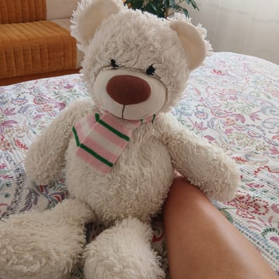 A plush, cream-colored teddy bear is lying on its side on a bed. The bear has a brown nose, black button eyes, and is wearing a striped scarf with pink and green lines. The bedspread is white with a colorful floral pattern in shades of pink, purple, green, and blue. To the right of the bear, a person's tanned leg is visible, resting on the bedspread. The leg is smooth and appears to have goosebumps. In the background, to the left of the bear, part of a green leafy plant is visible, along with a glimpse of a textured, orange sofa or cushion. The overall scene suggests a casual, indoor setting, possibly a bedroom or living room, during the daytime. The lighting is soft, indicating it could be morning or afternoon. The image is taken from a high angle, looking down at the bear and the leg.