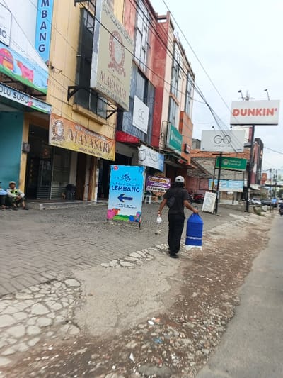 A street scene shows a man walking down a sidewalk, carrying a blue and white object. Buildings line the street with numerous signs, including "MAYASARI," "ROLU SUSU LEMBANG," "DUNKIN'," and "JAYA OPTIK." Other signs advertising various businesses are also visible. The sidewalk is paved with bricks, and there is visible street damage. Several people are sitting outside in the background.