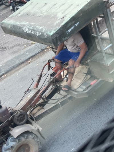 A man wearing beige shorts, a blue shirt, and black sandals is seated inside a large, olive-green metal structure attached to a tractor on a paved surface. The number "0942427986" is written on the side of the structure. The tractor has red handles and a small engine. The location is in M'Drắk, Vietnam.