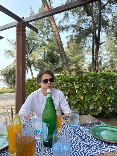 A person wearing sunglasses and a white shirt sits at a table outdoors, surrounded by lush greenery and palm trees. The table is covered with a patterned blue and white cloth, featuring plates, glasses filled with liquids, and a green water bottle. The scene suggests a relaxed outdoor dining experience in a tropical setting. A dark beam is visible in the foreground.
