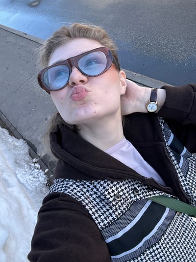A female individual is depicted in an upward-angled close-up selfie, situated outdoors in Новое Шигалеево, Russia. She wears large, brown-framed sunglasses with blue-tinted lenses and has her lips puckered. Her right hand is positioned at the back of her neck. On her left wrist, she wears a gold-toned watch with a dark brown strap and a white face featuring Roman numerals. She is dressed in a light pink top under a jacket with dark brown sleeves and hood, and a torso section displaying a black and white houndstooth pattern accented by horizontal black and white striped details. Metallic snap buttons are visible on the jacket's front. A dark green strap is visible across her chest. The background shows a dark, reflective paved surface on the right and a patch of dirty snow or ice on the ground to the left, indicating a cold environment.