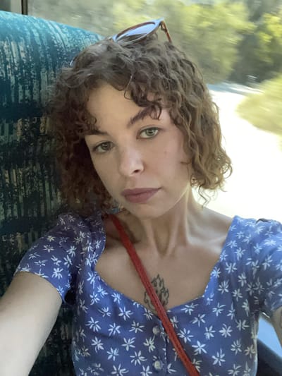 The media file shows a close-up, tilted selfie of a young woman. She has curly, reddish-brown hair and fair skin. Her eyes are green, and she has light pink lips. She is wearing a blue dress with a small white floral pattern and sunglasses pushed up on her head. A red strap, possibly from a purse or camera, is visible across her chest. A tattoo is partially visible on her chest. The background is blurred, suggesting movement, with streaks of green and yellow that could indicate trees or foliage seen through a window, possibly from a moving vehicle. The lighting suggests it is daytime. The overall mood appears casual and relaxed. The image is a selfie taken by the subject, showing her face and upper body. There is no visible text. The provided location context of Gornji Katun, Montenegro, suggests this might be a scene from her travels.