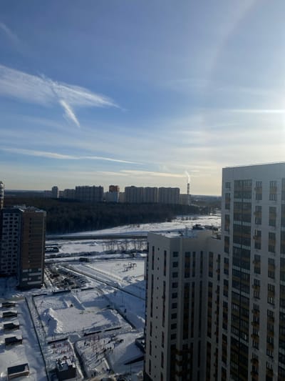 The image captures an expansive, elevated view of a snow-covered urban and forested landscape in Bittsa, Russia, under a clear, bright blue sky with scattered high-altitude clouds. The sun is visible in the upper right, casting a significant glare.
In the immediate foreground, two modern, multi-story residential buildings frame the lower left and right portions of the scene. The building on the right is light-toned with numerous dark-paned windows, while the building on the left is darker in color.
Directly below, a snow-covered ground plane reveals a residential courtyard or recreational area. Approximately 15-20 individuals, visible as small dark figures, are present in various clusters across the snow, suggesting general outdoor presence. The area contains snow-covered structures consistent with playground or outdoor fitness equipment. An adjacent snow-covered parking lot holds several parked vehicles.
The midground features a wide, snow-blanketed expanse, bisected horizontally by a multi-lane road or highway, partially cleared of snow. Beyond the road lies a dense, dark evergreen forest, likely Bittsa Forest, forming a prominent natural barrier.
In the distant background, a dense cluster of multi-story residential buildings extends across the horizon, forming a city skyline. To the far right of this background cityscape, a tall, slender industrial smokestack is visible, emitting a distinct plume of white smoke or steam vertically into the cold air. The overall environment indicates a clear winter day with pervasive snow cover. No specific interactions or detailed actions between individuals are discernible from this distance.