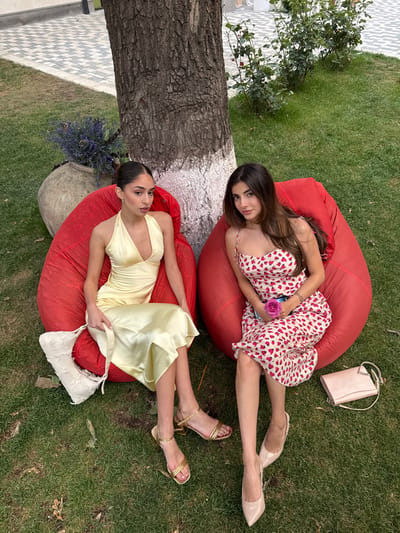 Two females are seated outdoors on red beanbag chairs, positioned on a manicured grass lawn in Nor Hachin, Armenia. A large tree trunk, painted white at its base, separates the subjects. The female on the left is seated on a red beanbag chair, wearing a light yellow satin midi-dress with a V-neck and halter straps, and gold-colored strappy sandals. She holds a cream-colored, textured shoulder bag on her lap. Her dark hair is pulled back. The female on the right is seated on a red beanbag chair, wearing a cream-colored midi-dress with red heart patterns and spaghetti straps, and light beige pointed-toe heels. She holds a single pink rose in her right hand. Her dark hair is long and flows over her shoulders. To the left of the scene, a large clay pot containing purple flowers is visible on the grass. To the right, a light pink clutch bag rests on the grass. In the background, a paved area with patterned tiles and green foliage is present. Both individuals are looking towards the camera.