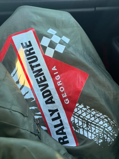 A close-up, high-angle shot shows a portion of a green jacket with graphics and text. The jacket has a zipper visible on the left side. A red and white sticker or graphic is layered on the jacket. The text on this graphic reads "RALLY ADVENTURE GEORGIA" in black and white lettering. Behind the text, a checkered flag pattern and a tire track graphic are visible on the green fabric. The lighting suggests this is an indoor or shaded outdoor setting, with a reflection of light on the sticker. The overall scene is static and focused on the jacket's design. No specific location within the city of Samtsevrisi, Georgia can be determined from the image.