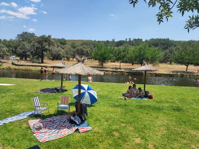 The image depicts a sunny day at a natural swimming area located in Cadalso, Spain. A group of people are enjoying the water and the grassy bank. In the foreground, several beach towels are spread out on the green lawn, with a couple of folding chairs set up. One person is seated in a striped chair, while another is in a brown chair. Nearby, a blue and white umbrella provides shade.
In the middle ground, several people are visible in the water, some swimming and others wading. On the grassy bank near the water's edge, a small group of people are sitting together.
The background shows a serene landscape with trees and hills. The water appears calm, reflecting the clear blue sky. The overall scene conveys a relaxed and pleasant atmosphere, characteristic of a summer day spent outdoors.