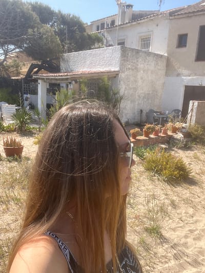 The image, taken outdoors in daylight, features a woman with long, wavy brown hair partially visible. The location appears to be La Marina De Creixell, Spain, evidenced by the style of residential buildings in the background. The background includes two buildings with white exteriors and brown window shutters. Several potted plants are arranged against one of the buildings. The ground is covered in sand with sparse vegetation. The woman is wearing sunglasses and a garment with the word "EMPORIO" visible. She seems to be either lying down or reclining, with her hair spread out on the sandy ground.