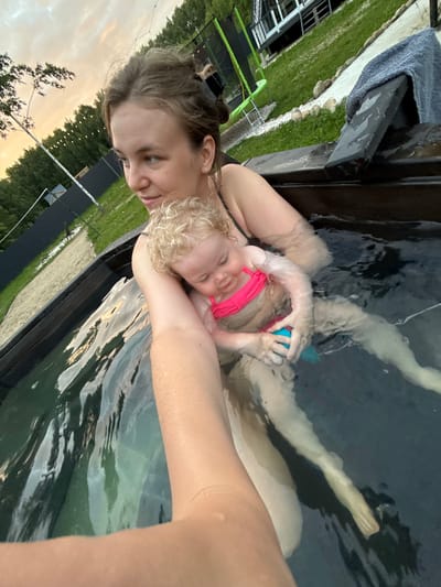 This media file is a selfie of two people, a woman and a child, in a hot tub. The woman is holding the child, who is wearing a pink swimsuit and has blonde curly hair. The child appears to be asleep or resting on the woman's chest. The water in the hot tub is dark, and the surroundings are visible in the background, including grass, trees, and some outdoor lighting. The image is oriented sideways, suggesting it was taken with a phone and possibly rotated for viewing. The time of day appears to be late afternoon or early evening, as indicated by the warm, soft light and the sky in the background. The overall mood seems peaceful and intimate, capturing a moment of relaxation and connection between the two individuals. The location is identified as Ryzhkovo, Russia, but there are no explicit visual cues in the image to confirm this. There is no visible text in the image.