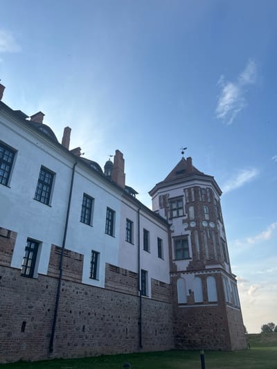 The image shows the Mir Castle Complex, a UNESCO World Heritage site located in the village of Mir, Korelichsky District, Grodno Region, Belarus. The photograph is taken from a low angle, looking up at a section of the castle complex under a bright, partly cloudy blue sky.
The main subjects are the castle walls and a prominent tower. The wall in the foreground is constructed with large, rough stones at the base, transitioning to a brickwork section above, topped by a smooth, white plastered facade. Several dark-framed windows are visible on the white upper section. The tower to the right features a more intricate design, with red brickwork and white decorative elements. It is octagonal in shape and has multiple windows. A weather vane is visible at the very top of the tower.
The environment is an outdoor setting with green grass visible at the bottom of the frame, suggesting it is a park or grounds surrounding the castle. The sky is a clear blue with scattered wispy clouds and a few small, distant birds. The lighting suggests it is daytime, possibly late afternoon due to the angle of the sun which casts a slight shadow on the buildings. There are no people visible in the image.