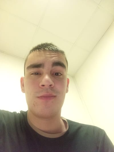 A close-up selfie shows a young man, likely in his late teens or early twenties, looking directly into the camera. He has short, dark, textured hair, pronounced dark eyebrows, and brown eyes. A faint shadow of stubble can be seen on his chin and upper lip. He is wearing a dark t-shirt with a lighter-colored, possibly beige or light brown, trim along the collar.

The image is framed from a slightly low angle, capturing the man's head and upper torso. The background primarily features a plain, light-colored (off-white or cream) ceiling with visible grid lines, suggesting acoustic or modular ceiling tiles. A section of a light-colored wall, forming a corner with the ceiling, is also visible on the right. The lighting is bright and artificial, consistent with overhead indoor illumination.

The simple, functional nature of the ceiling and wall indicates an indoor, possibly utilitarian, setting. This environment aligns with the context of a camp, such as the Tecnimont camp in Russia. The man's expression is neutral to mildly pleasant, with a slight upturn at the corners of his lips. No other subjects, distinct objects, or text are visible in the frame.